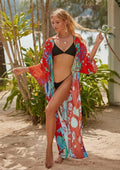Woman in a colorful floral kimono and black bikini standing on a sandy beach with plants in the background.
